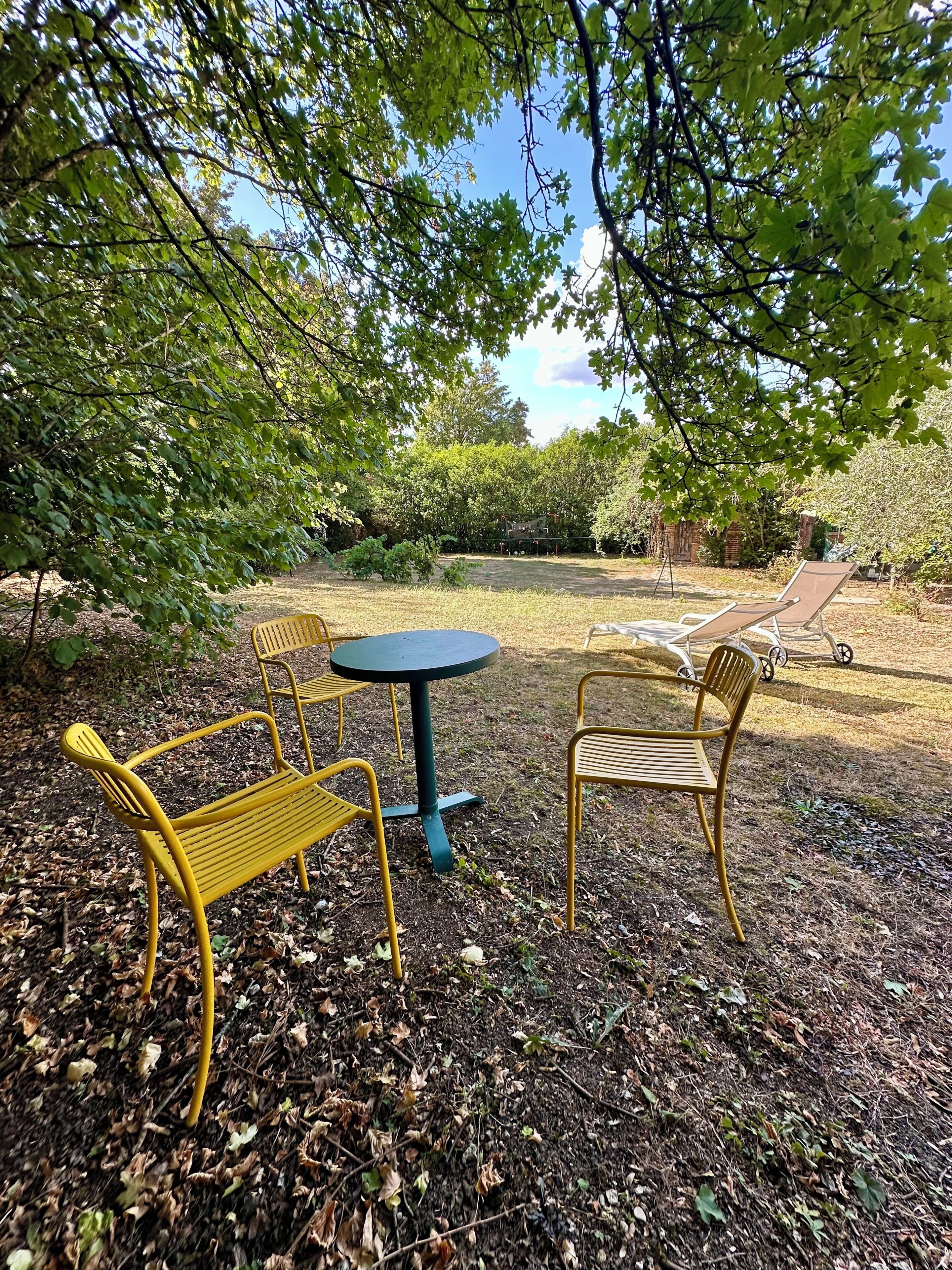 Un espace extérieur aménagé avec des chaises en métal jaune et une table ronde, entouré d’arbres et d’un jardin.