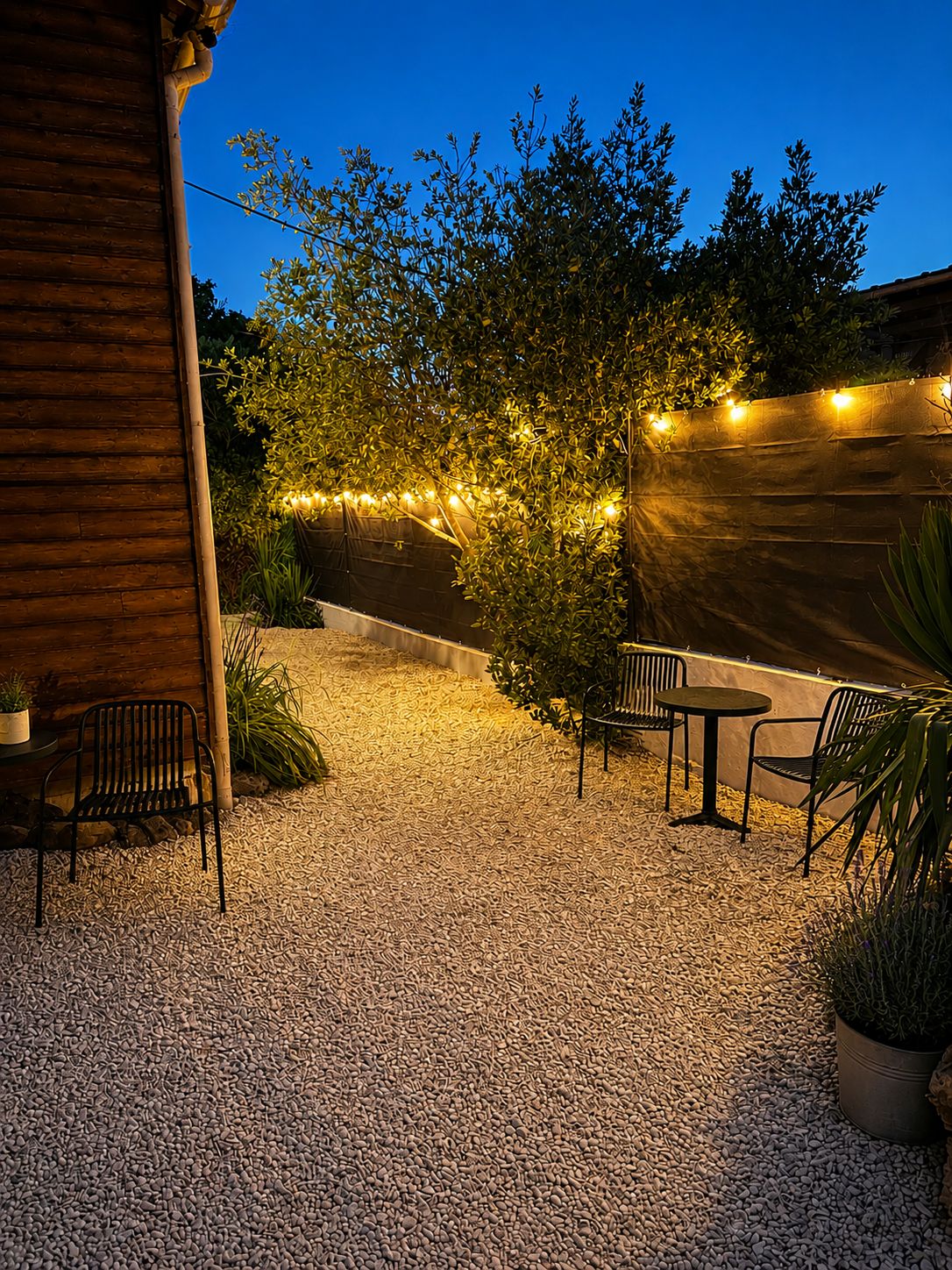 An outdoor patio area with a gravel floor, metal chairs, a small table, and string lights.