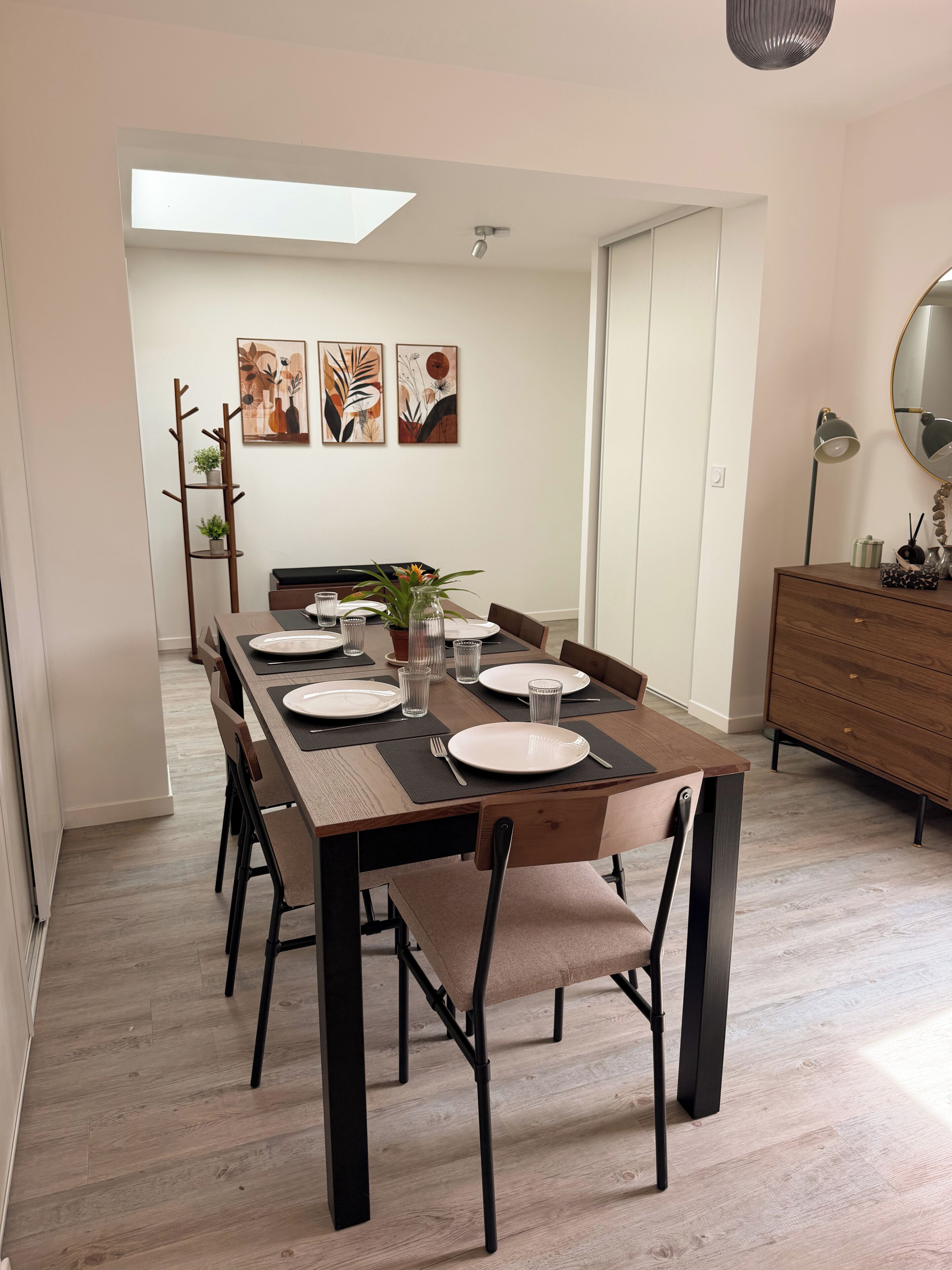 Uma área de refeições com uma mesa de madeira comprida posta para quatro pessoas, com cadeiras ao redor e uma luminária moderna em um aparador.