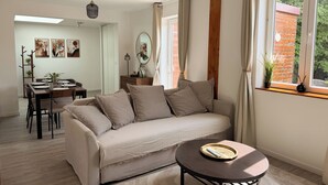 Een moderne woonkamer met een bank, salontafel, eettafel en stoelen. Er is een raam met gordijnen, een plant op de vensterbank en kunstwerken aan de muren.