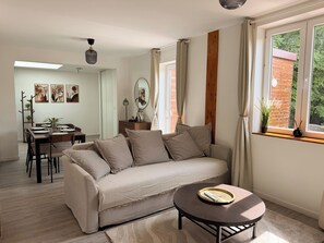 En moderne stue med sofa, sofabord, spisebord og stole. Der er et vindue med gardiner, en plante på vindueskarmen og kunst på væggene.