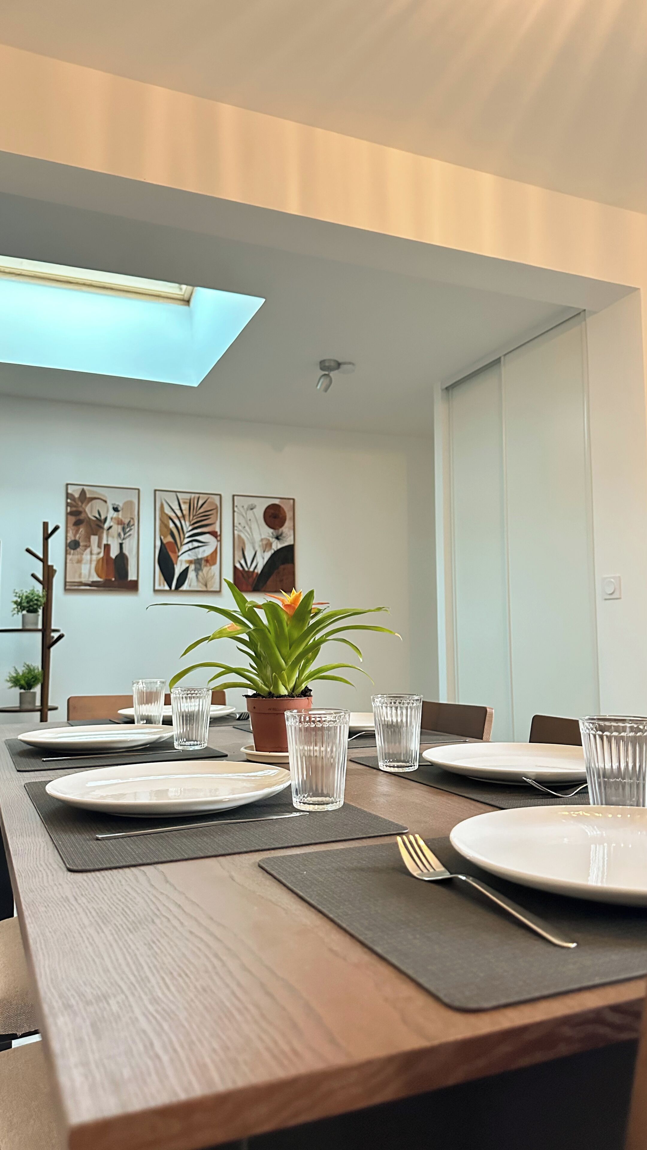 Un coin repas avec une longue table dressée pour quatre personnes, agrémentée de beaux verres, d’assiettes et de couverts. Deux plantes en pot sont posées sur la table, et la pièce présente un design moderne avec des œuvres d’art abstraites accrochées aux murs.