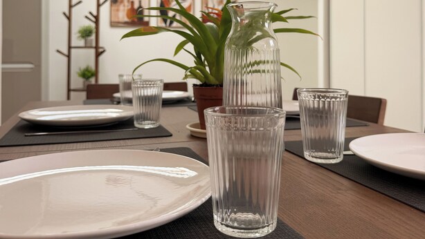 Een eettafel gedekt met borden, glazen en een vaas, met een plant en kunstwerk op de achtergrond.