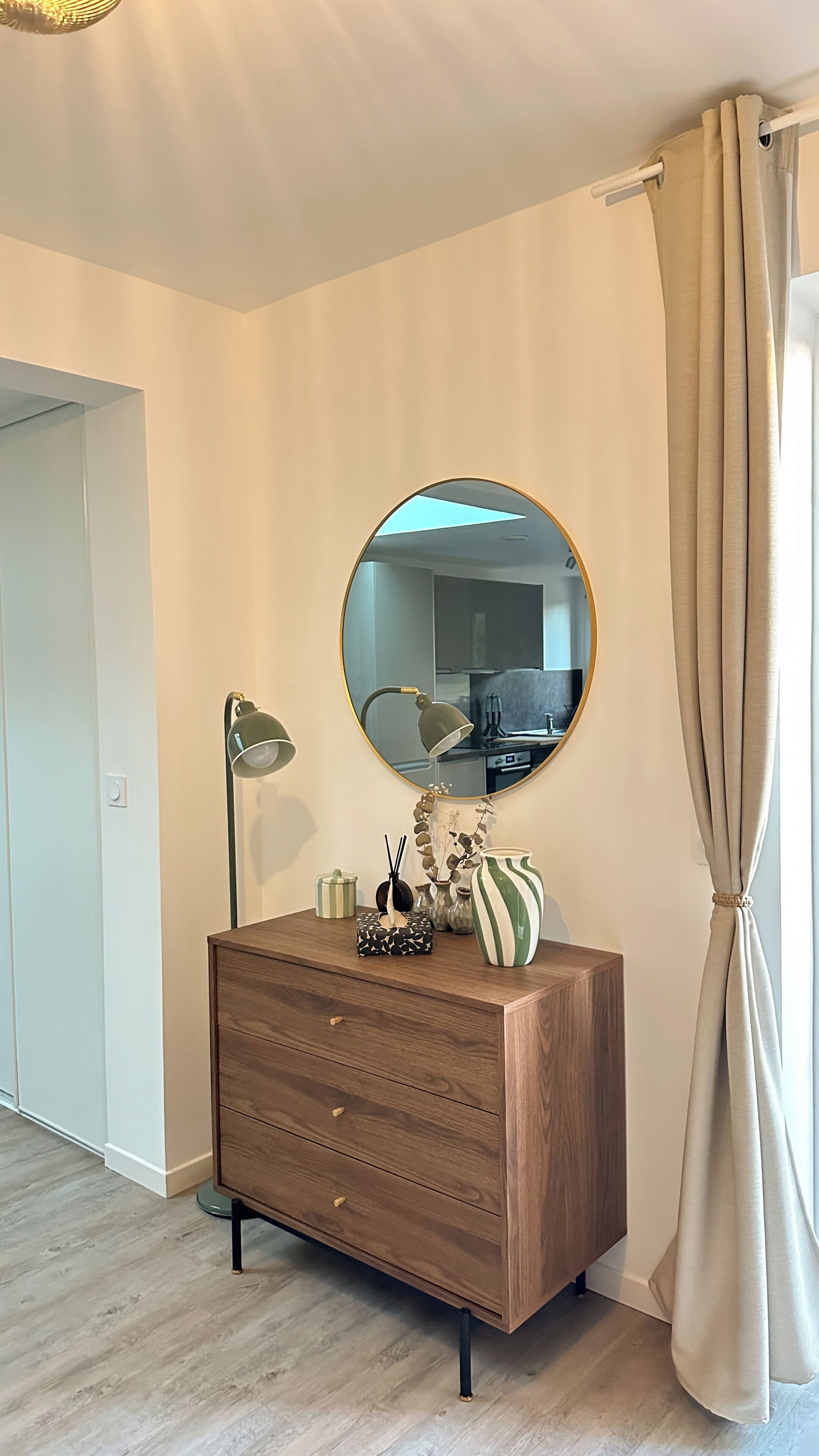 A room with a wooden dresser, a round mirror, a lamp, and a window with curtains.