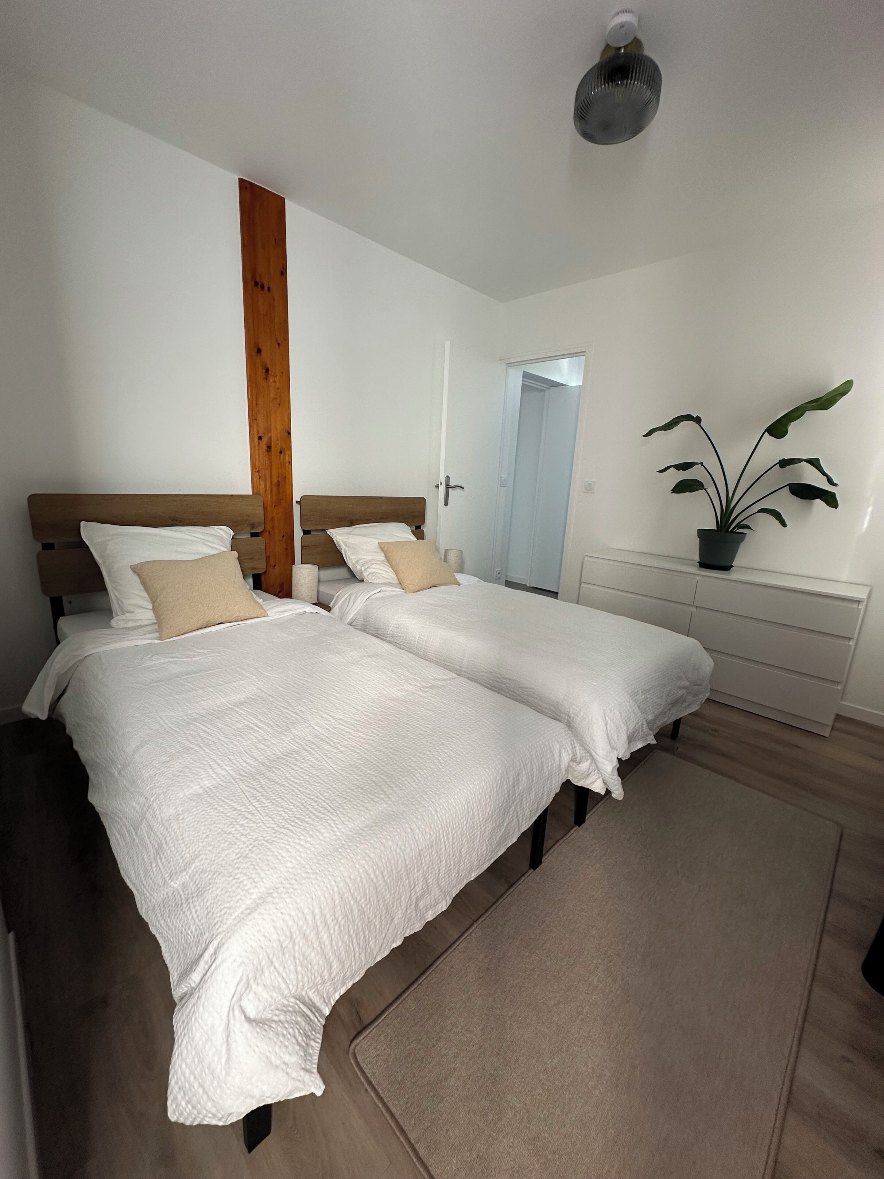 Quarto com duas camas, uma parede de destaque em madeira, um vaso de planta e uma cômoda branca.
