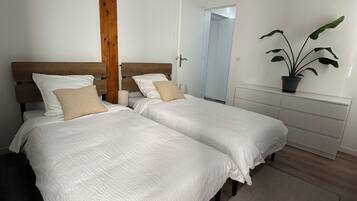 Quarto com duas camas, uma parede de destaque em madeira, um vaso de planta e uma cômoda branca.