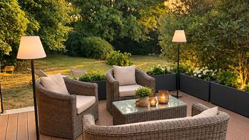 Un patio al aire libre con mobiliario de mimbre, incluyendo un sofá y sillas, una mesa de centro con superficie de vidrio y dos lámparas de pie.