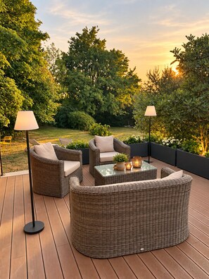 Un patio al aire libre con mobiliario de mimbre, incluyendo un sofá y sillas, una mesa de centro con superficie de vidrio y dos lámparas de pie.