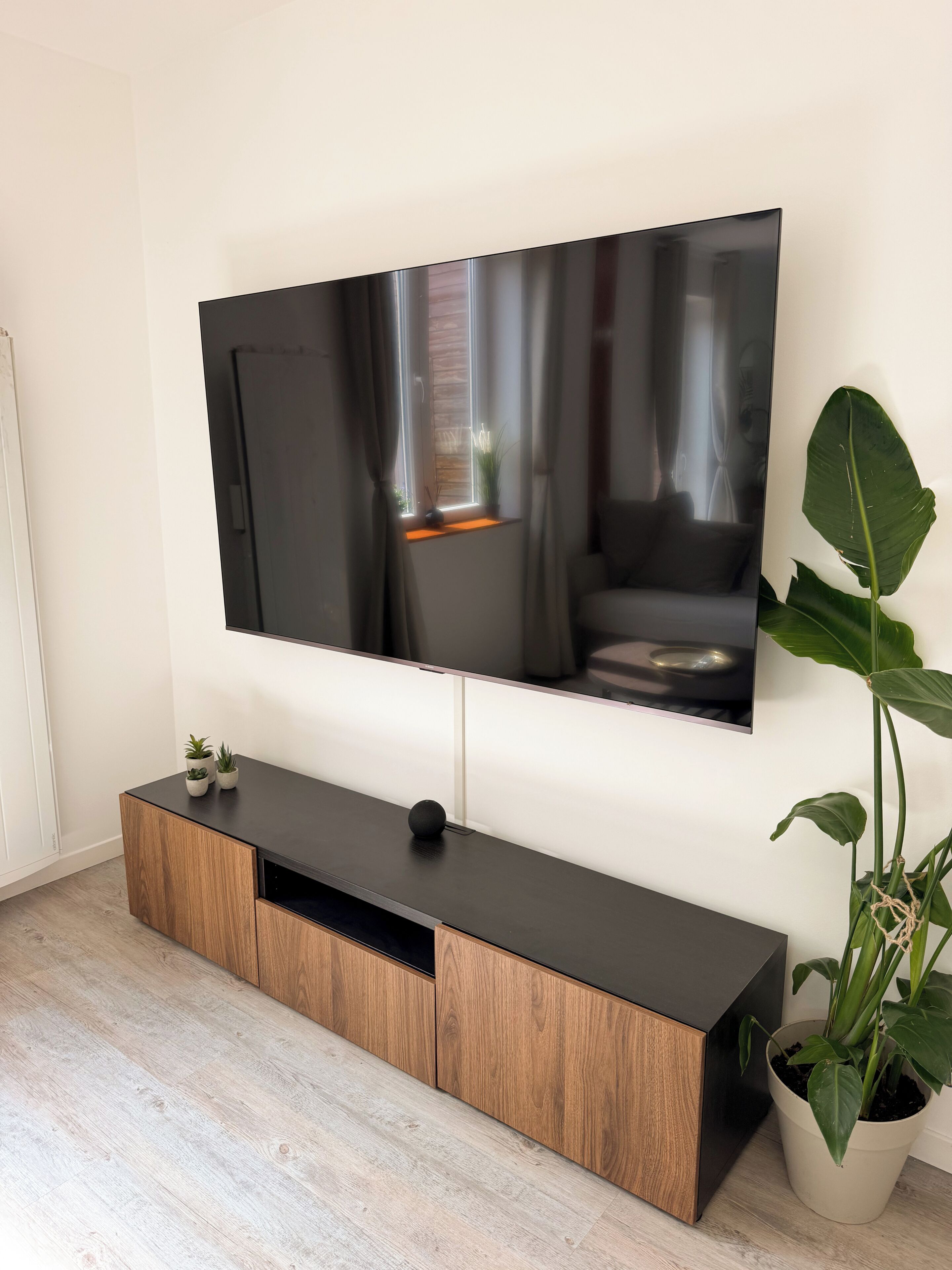 Een flatscreen televisie hangt aan een muur, boven een houten tv-meubel. Daarbij staat een potplant.