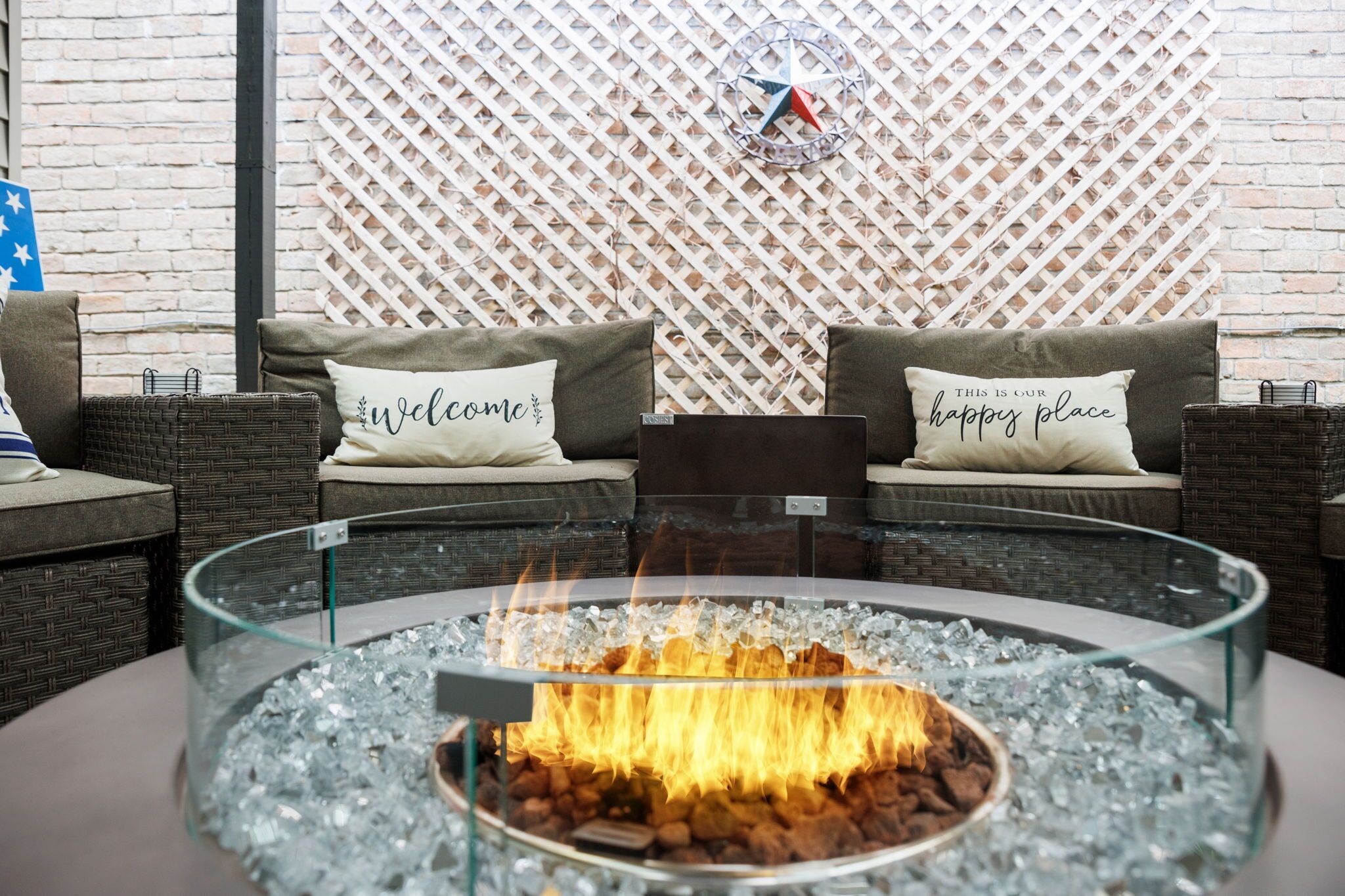 A glass-top fire pit with a fire burning, surrounded by cushions with 'welcome' and 'happy place' text, against a brick wall with a lattice pattern.