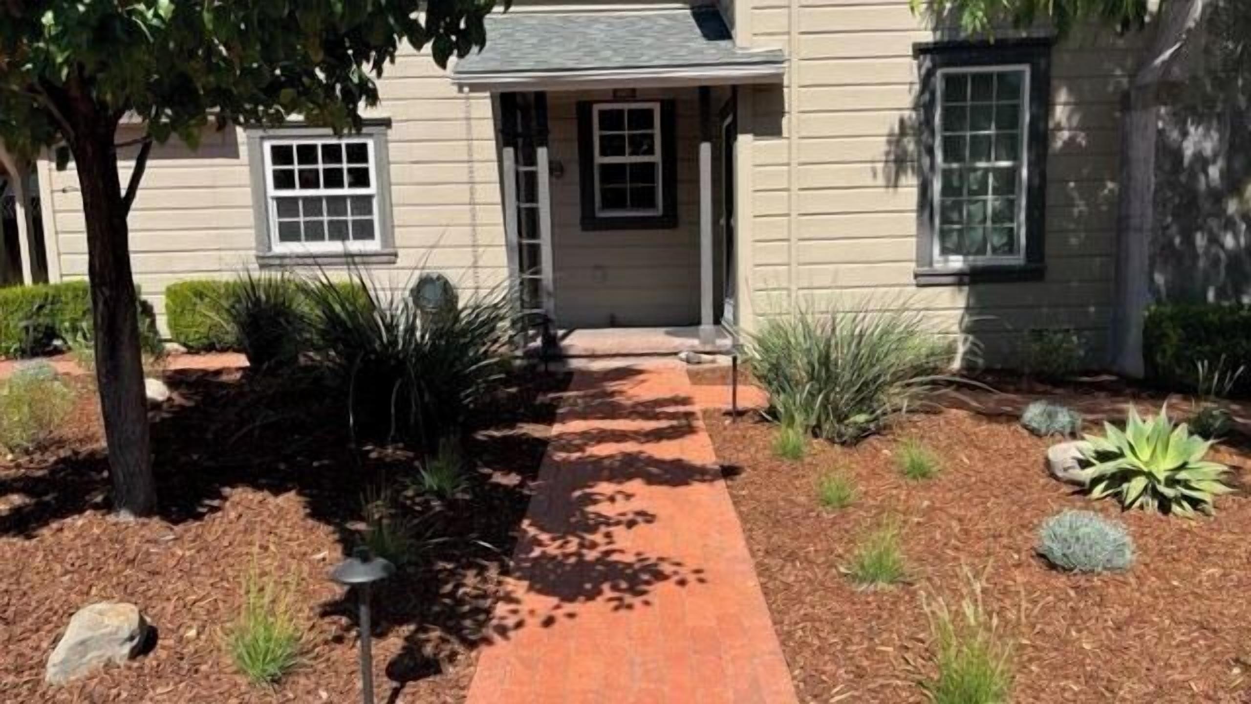 A house with a brick pathway leading to the front door, surrounded by landscaped garden beds and a variety of plants.
