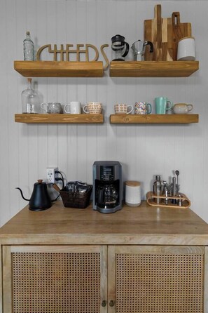 Una cucina con scaffali in legno che contengono tazze e utensili da cucina, una macchina per il caffè e un armadietto con pannello frontale in vimini.