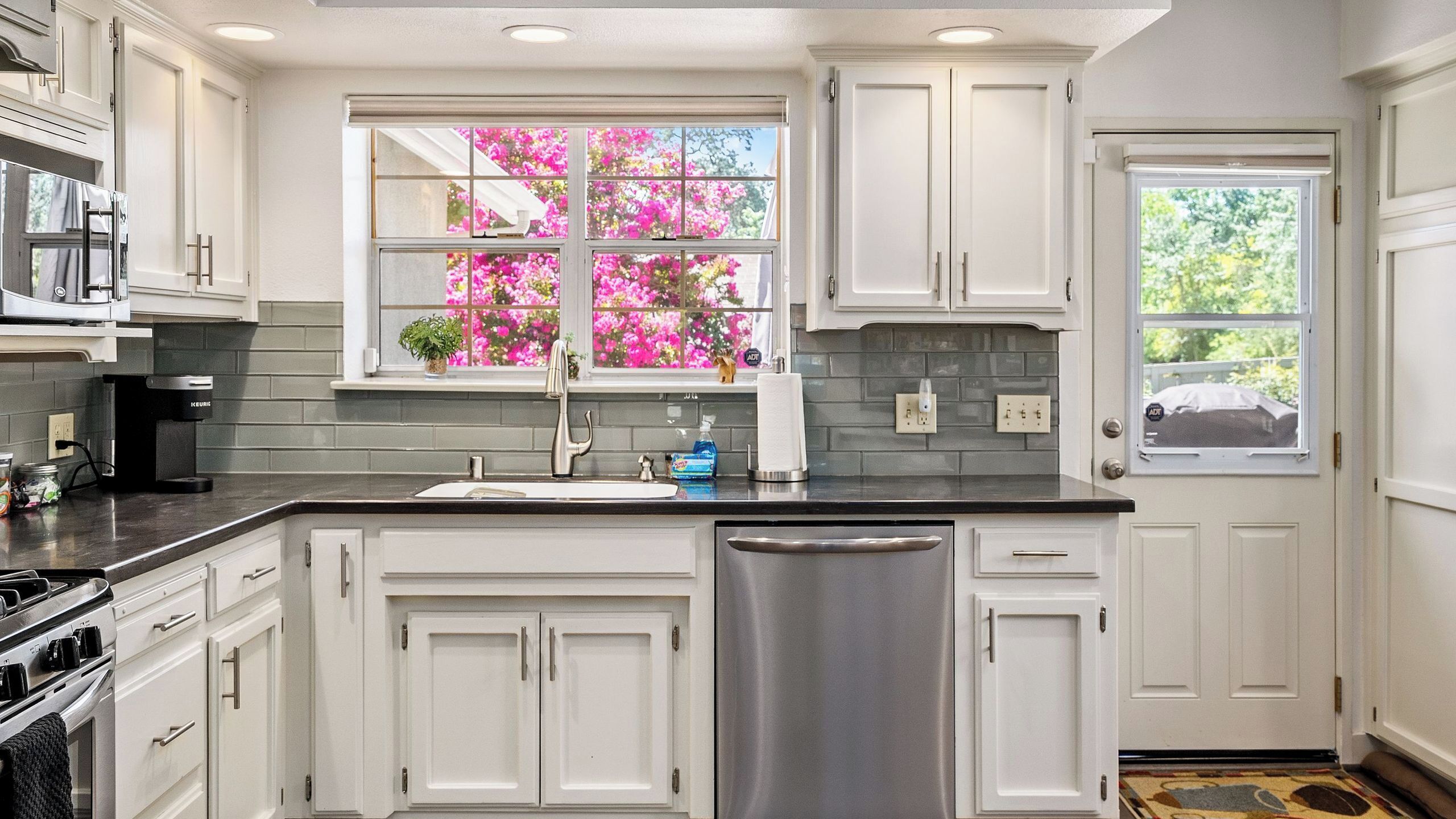 A kitchen with white cabinets, a stainless steel dishwasher, and a window with pink flowers.