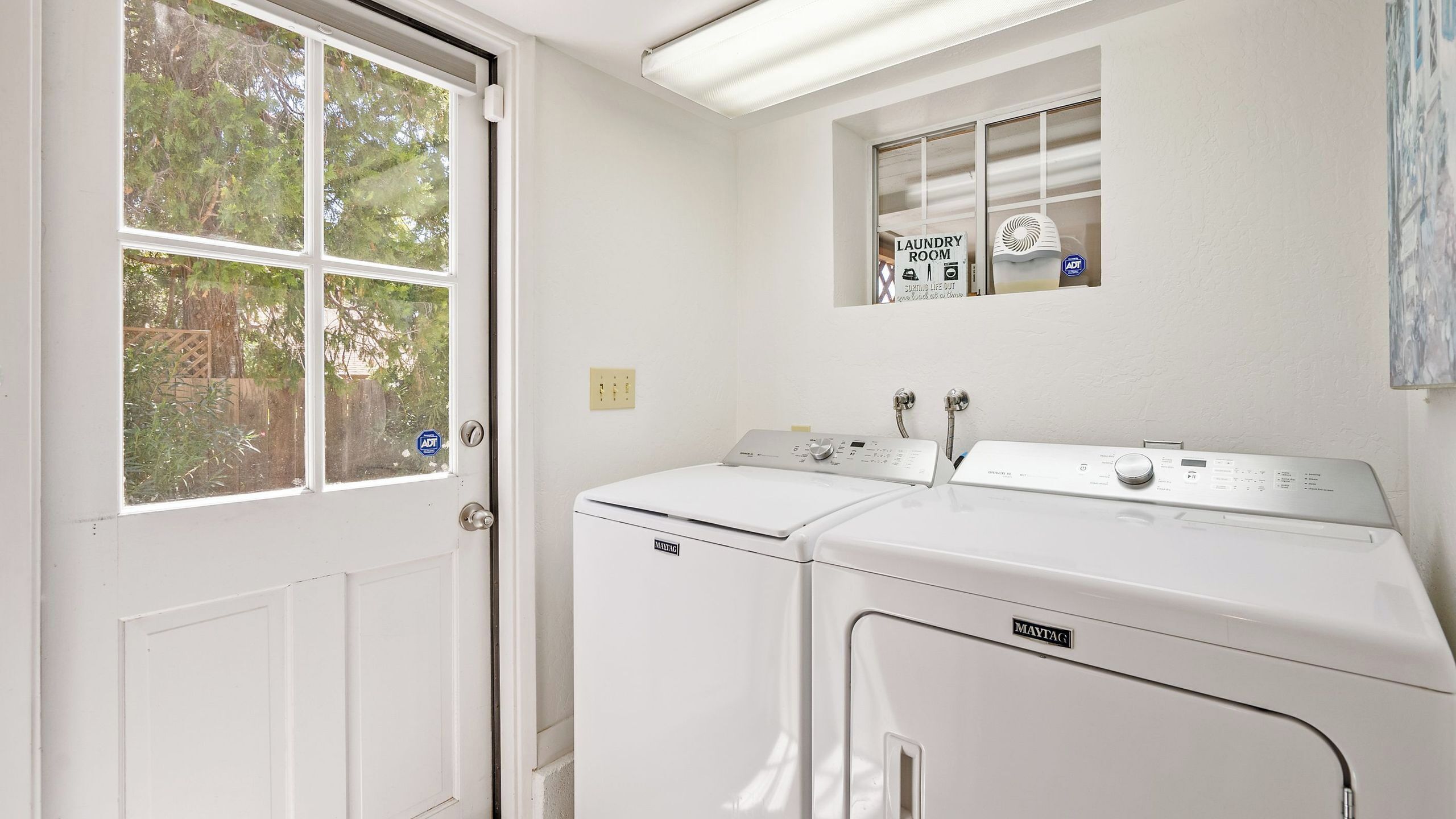 A laundry room with two white Maytag washers and dryers, a window with a view of trees, and a sign indicating it's a laundry room.