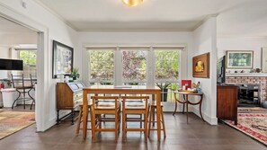 A dining area with a wooden table and chairs, a kitchen with a brick wall, and large windows with a view of greenery. - Chateau Paso by Avantstay Pool Table + Private Deck Walk to Downtown Paso (Paso Robles)