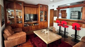 Sala de estar com sofá, mesa de centro e televisão. Há armários de madeira e uma área de cozinha com frigorífico.