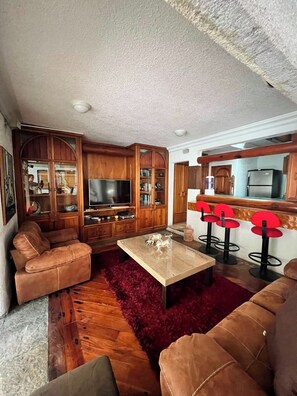 Una sala de estar con un sofá, una mesa de centro y un televisor. Se observan armarios de madera y una zona de cocina con un refrigerador.