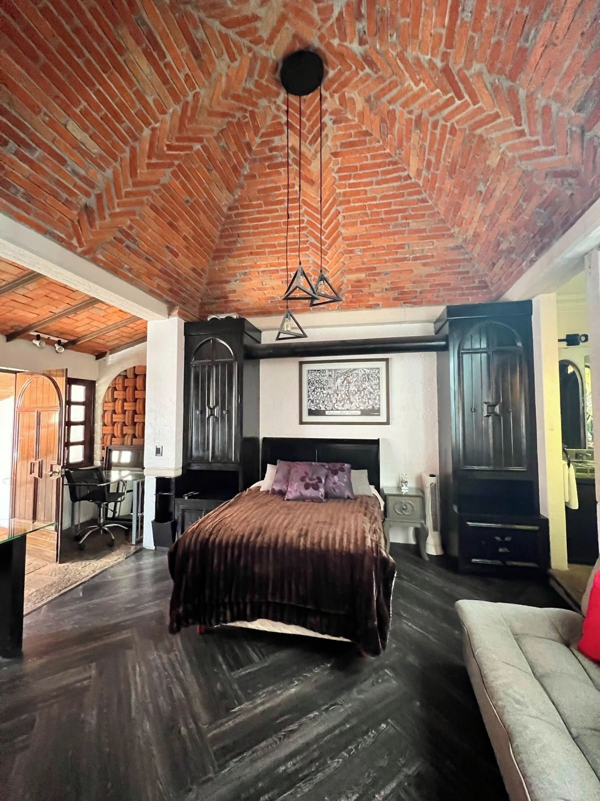 Un dormitorio con techo de ladrillo, una cama con colcha marrón oscuro, un escritorio con silla y un ventanal grande.