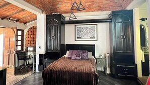 Um quarto com teto de tijolos, uma cama com colcha marrom escura, uma escrivaninha com cadeira e uma janela grande.
