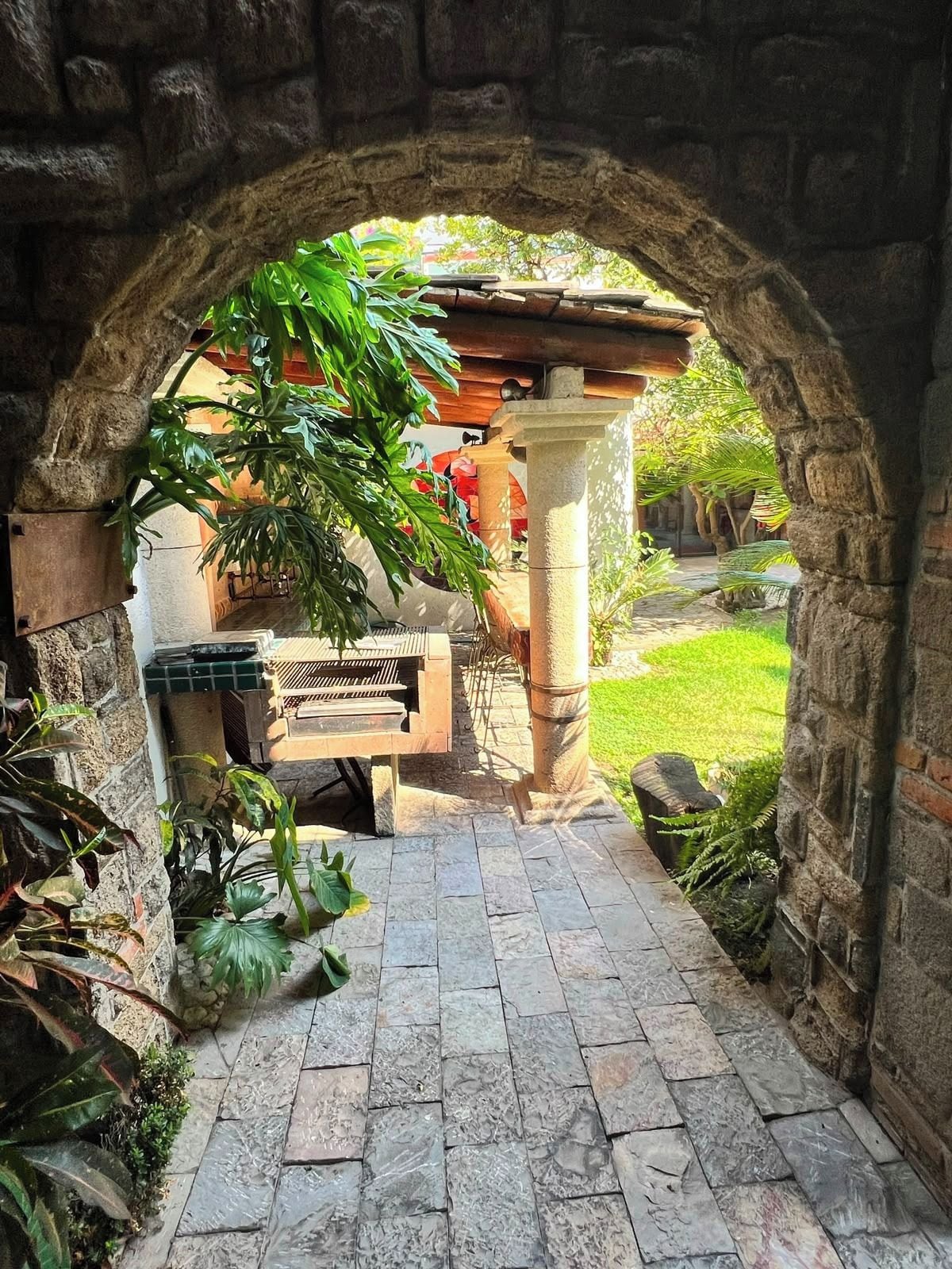 An outdoor stone archway leading to a patio area with a bench and a table, surrounded by greenery.