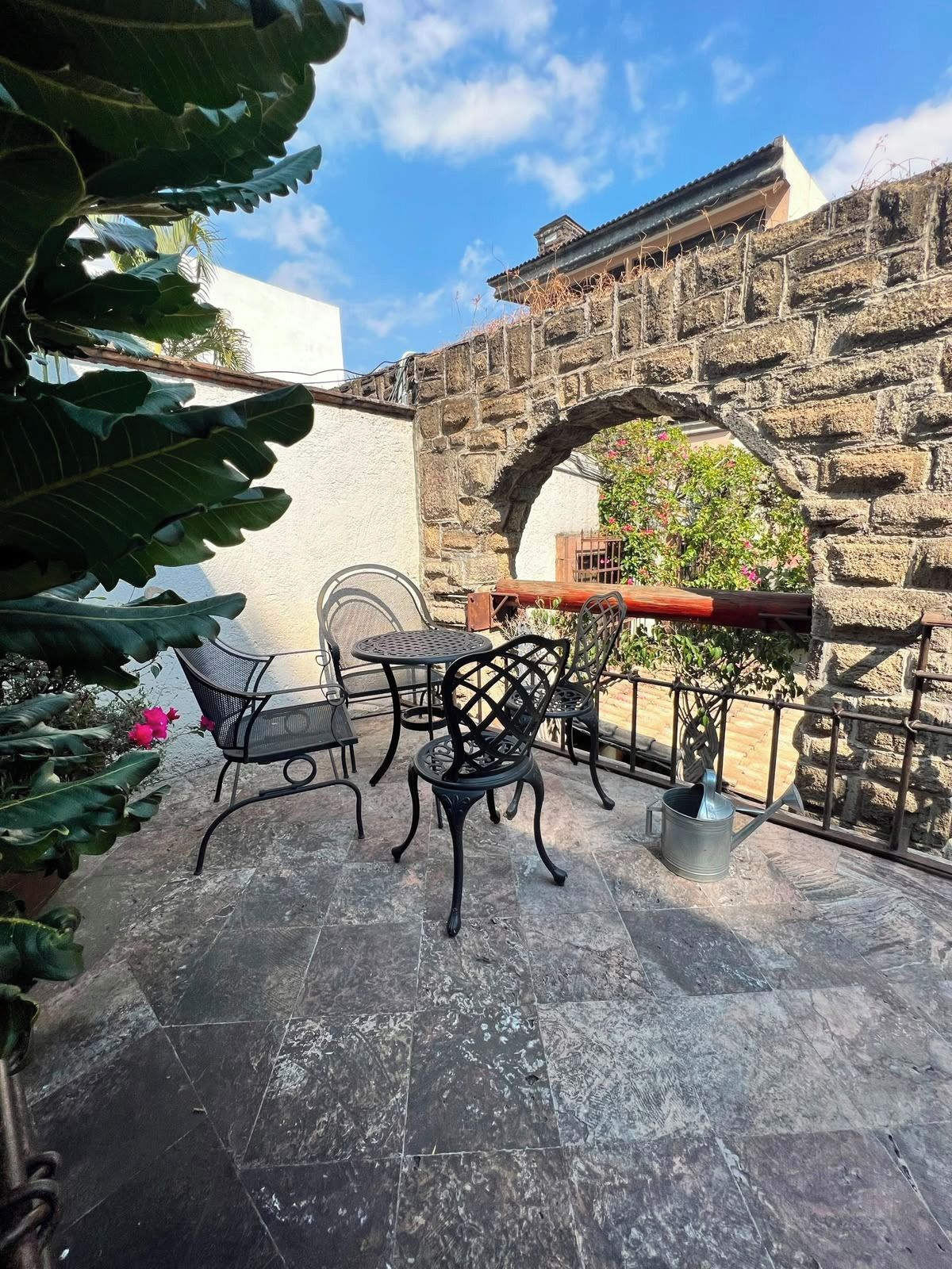 Un patio exterior con suelo de piedra, sillas de metal, una mesa y un muro de piedra con un arco.