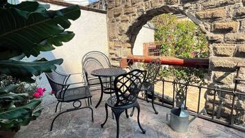 Un patio exterior con suelo de piedra, sillas de metal, una mesa y un muro de piedra con un arco.