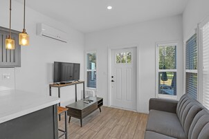 A modern living room with a flat-screen TV, a sofa, a dining table, and a kitchen area with a built-in oven.