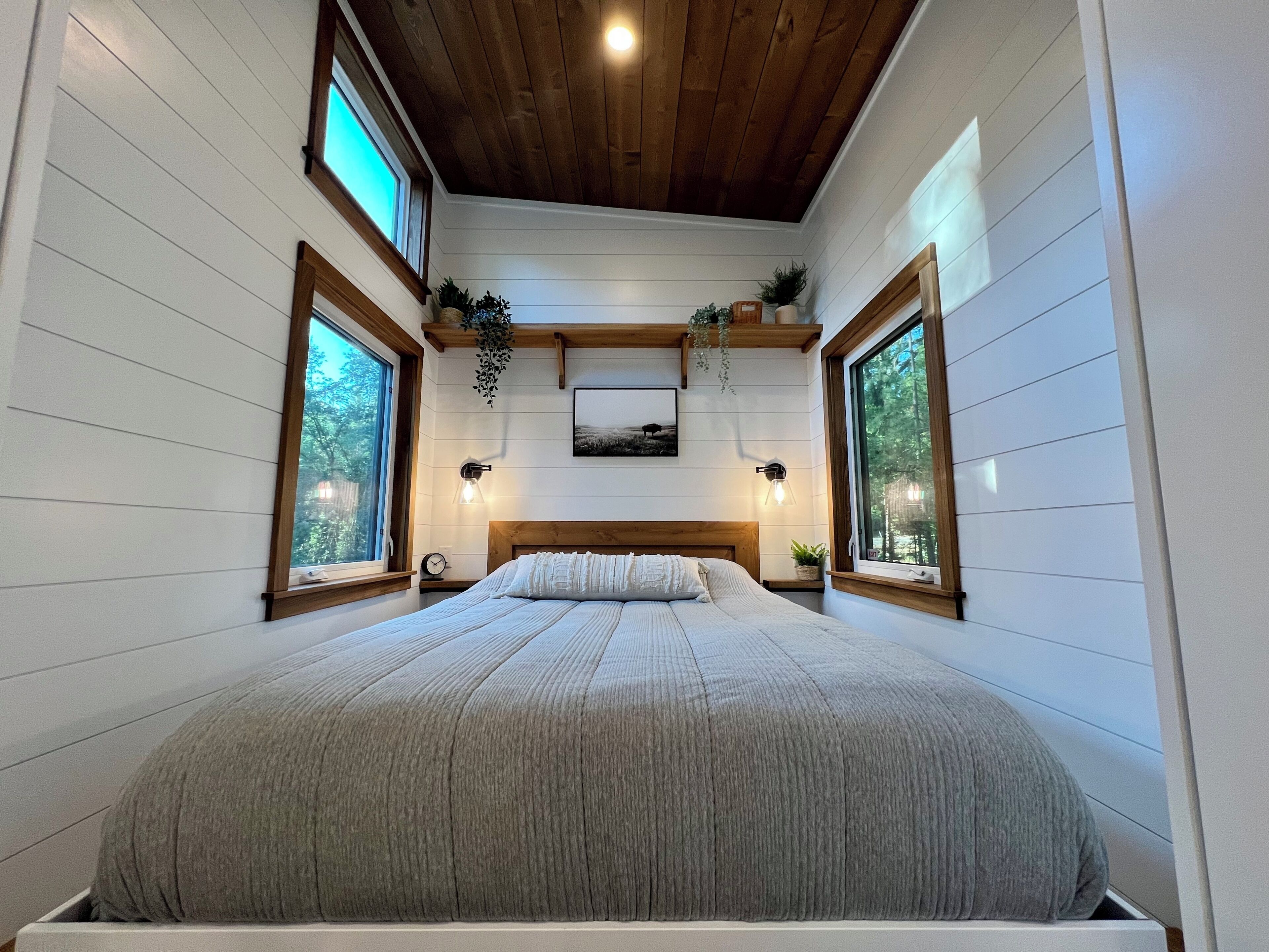 A bedroom with a large bed, wooden ceiling beams, and two windows with plants.