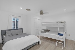 A bedroom with a bunk bed, a single bed, a window with curtains, and a door. - Tiny House Village (Groveland)