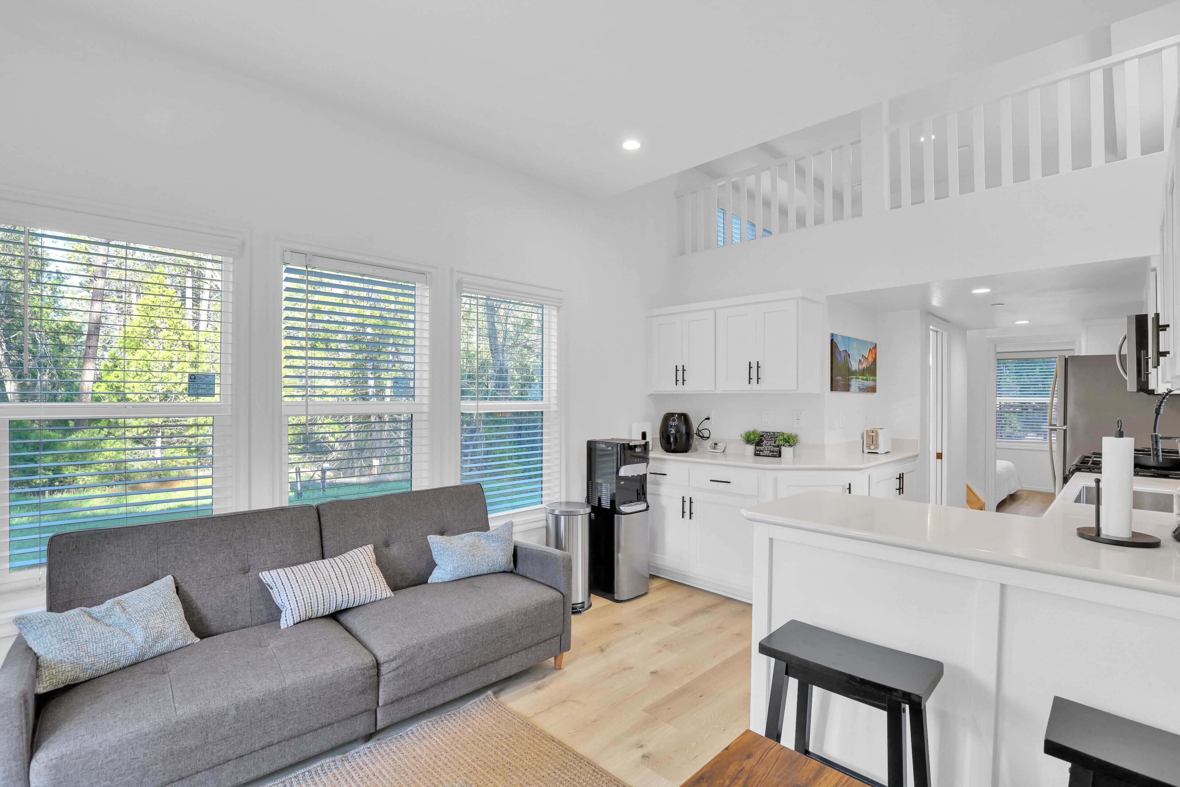 A modern apartment with a grey sofa, a kitchen with white cabinets, and large windows with blinds.