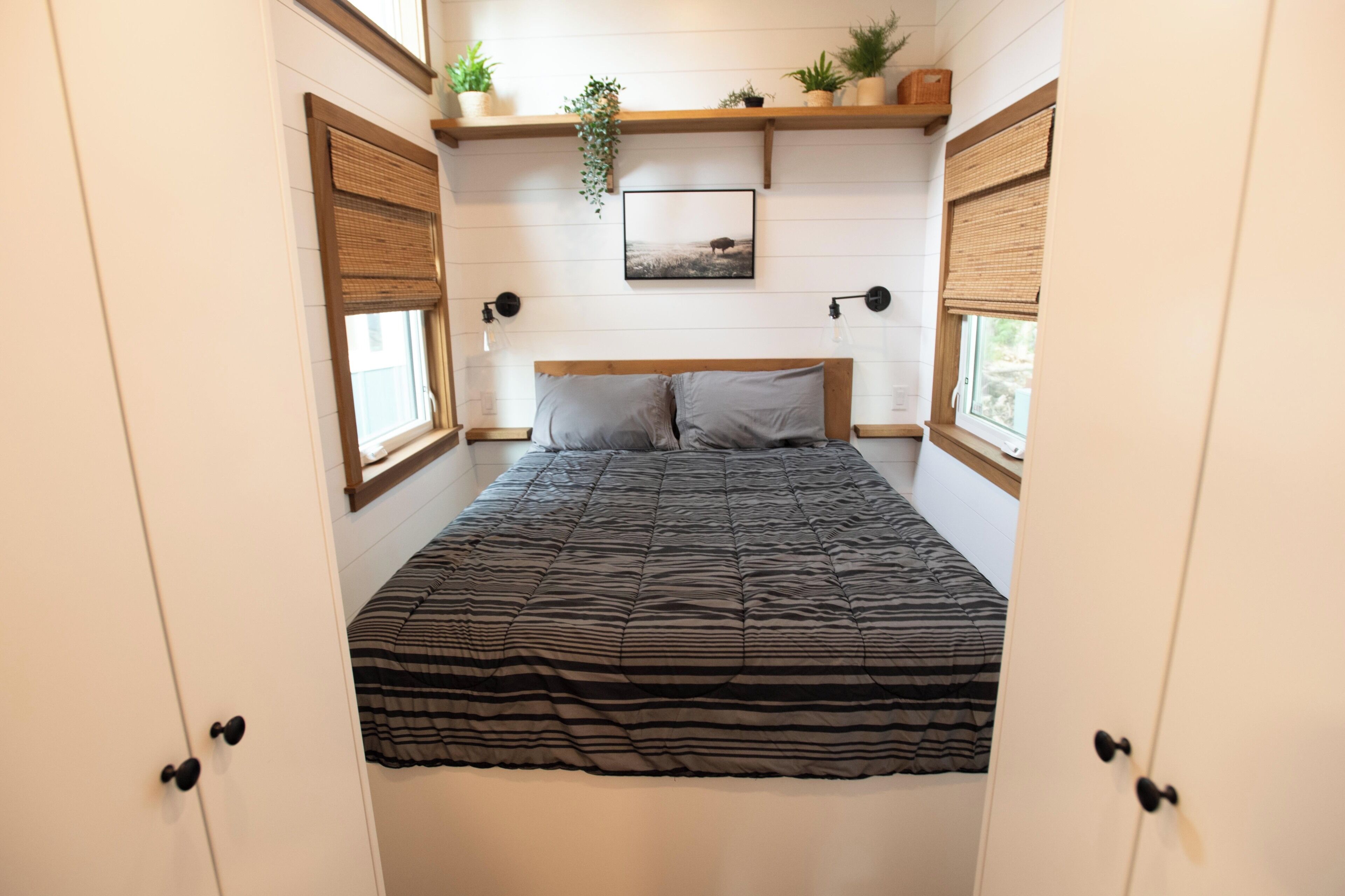 A bedroom with a bed, two bedside tables, a wall-mounted shelf with plants, and a window with a view.