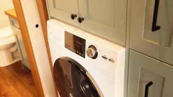 A compact, modern laundry room with a front-loading washing machine, wooden flooring, and light-colored cabinetry.