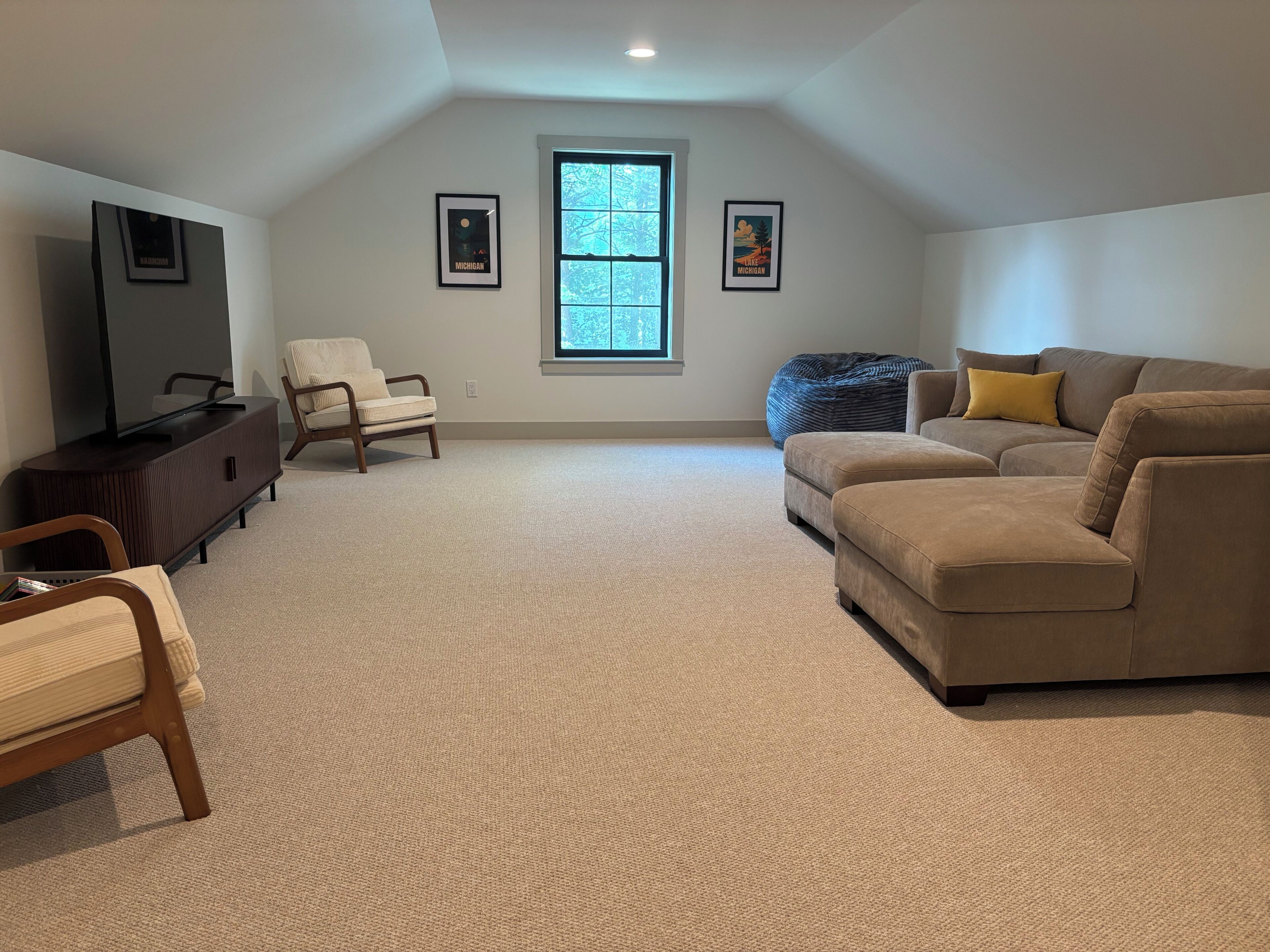 A spacious living room with a large window, a flat-screen TV, a beige sofa, a wooden chair, and a bean bag.
