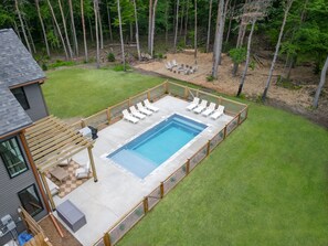 Ein rechteckiger Swimmingpool mit Holzterrasse und Liegestühlen, umgeben von einem Holzzaun, mit einer Rasenfläche und Bäumen im Hintergrund.