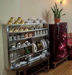 A kitchen area with a metal shelf unit holding various kitchenware, a refrigerator with a floral design, and a potted plant on a wooden table.