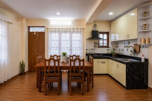 A dining area with a wooden table and chairs, a kitchen with a black countertop and yellow cabinets, and a window with sheer curtains. - Serenova Apartment,  Modern 2BHK, Valley View (Kathmandu)