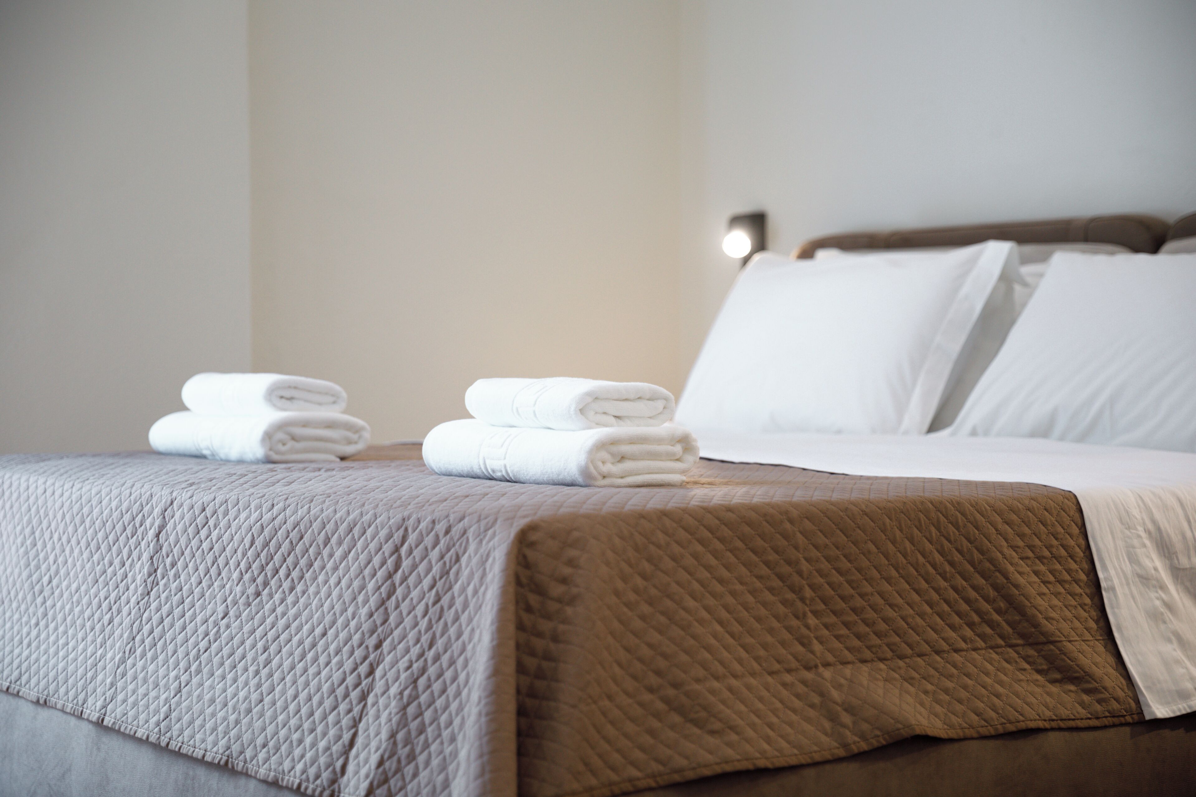 A neatly made bed with a quilted bedspread, two folded towels, and a headboard with a light fixture.