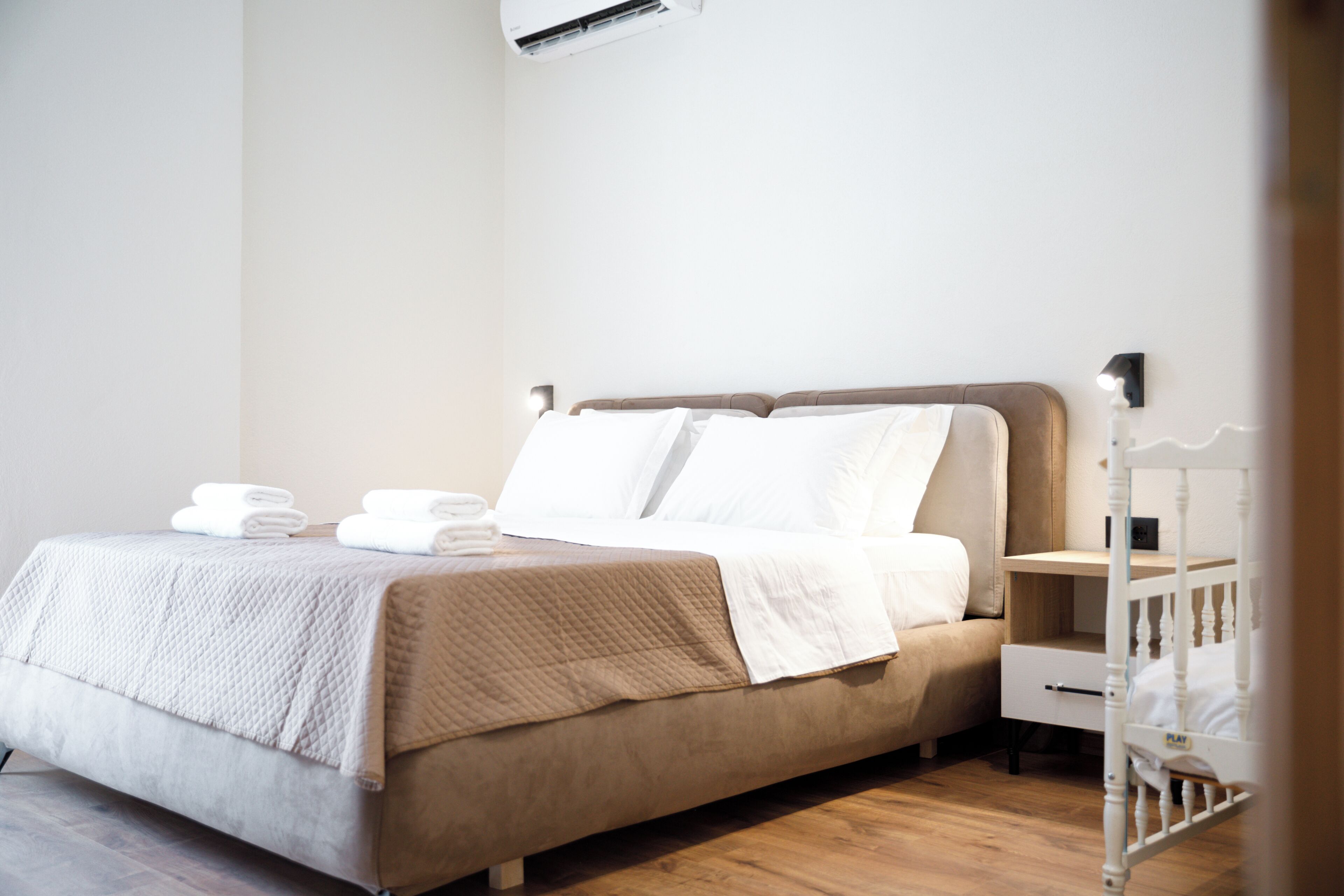 Ein ordentlich bezogenes Bett mit weißen Leinentüchern und einer beige farbenen gesteppten Decke, ein Nachttisch mit Lampe sowie ein weißes Babybett in der Ecke.