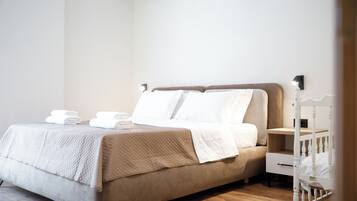 Ein ordentlich bezogenes Bett mit weißen Leinentüchern und einer beige farbenen gesteppten Decke, ein Nachttisch mit Lampe sowie ein weißes Babybett in der Ecke.