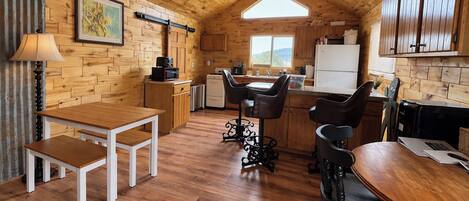 A cozy kitchen with wooden walls and a sloped ceiling, featuring a dining table, bar stools, and a refrigerator.