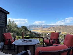 An outdoor patio with a fire pit table, red cushioned chairs, and a view of mountains and a lake.