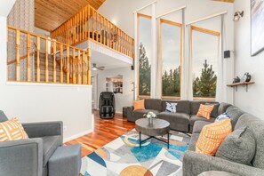 A spacious living area with a grey sofa set, a massage chair, and a coffee table. A wooden staircase with a railing leads to an upper level.