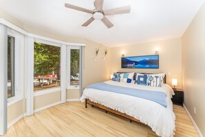 A bedroom with a bed, a ceiling fan, a window with a view of the outdoors, and a painting on the wall.