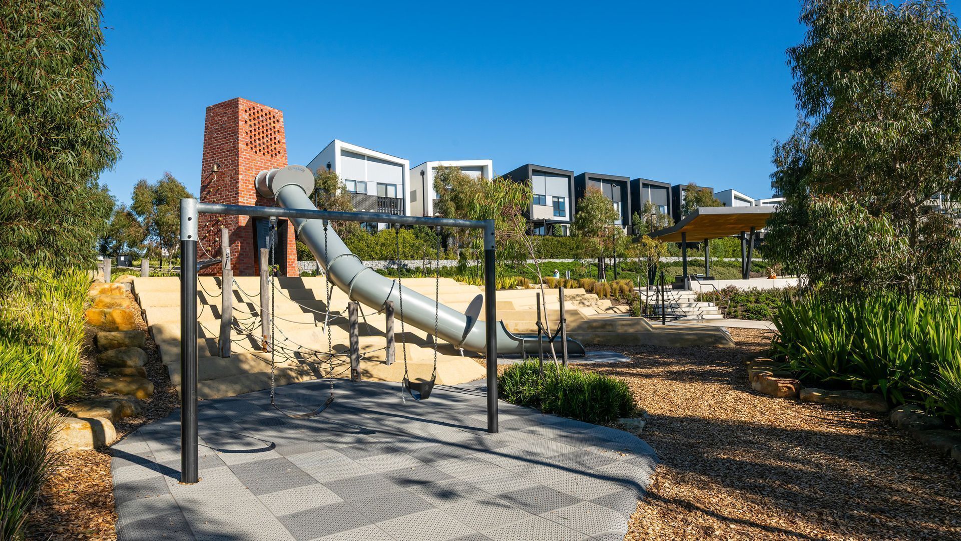 An outdoor playground with a slide and swings, surrounded by landscaped areas and modern buildings in the background.