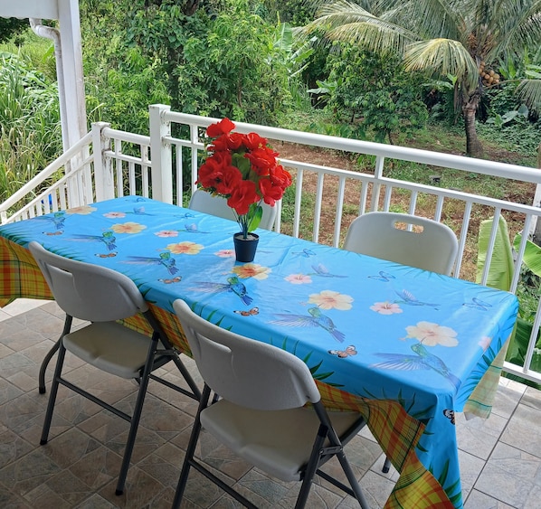 A table with a floral tablecloth, two chairs, and a potted plant with red flowers on a balcony with a white railing.