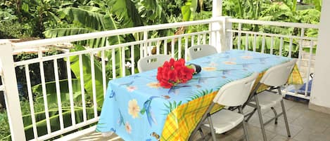 A covered outdoor dining area with a table set for two, surrounded by a white railing, and lush tropical foliage in the background.
