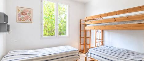 Une chambre avec deux lits superposés, une fenêtre donnant sur un paysage verdoyant, un luminaire suspendu et un tableau encadré au mur.