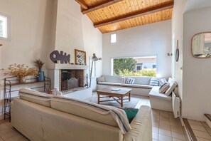 A living room with a fireplace, a sofa, a coffee table, and a window with a view of greenery. - La Linière - Proche plage - Petfriendly (Noirmoutier-en-l'Île)