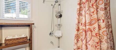 A bathroom with a shower, a bathtub, a window with blinds, and a patterned curtain.