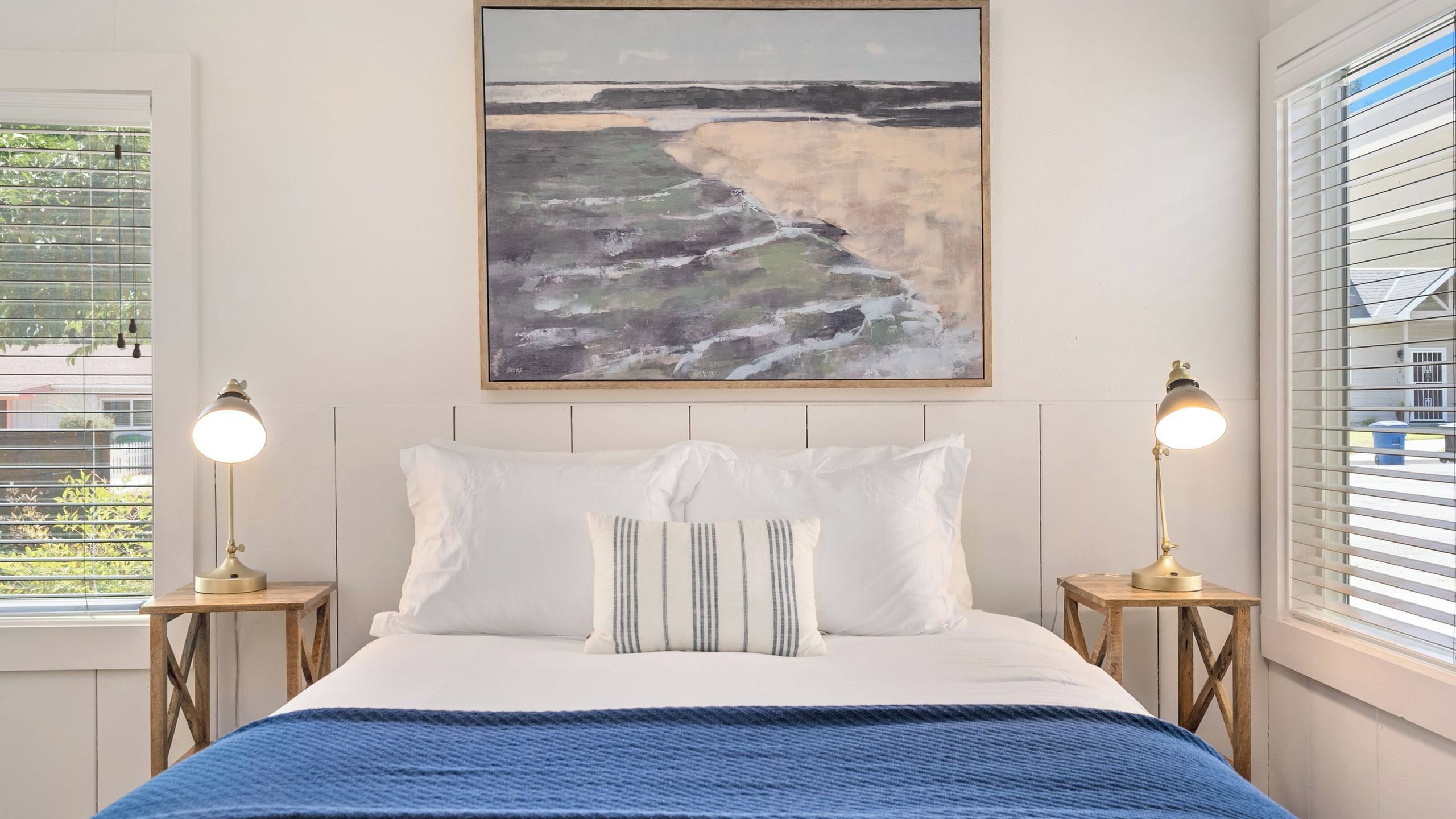 A bedroom with a bed, two bedside lamps, a painting of a beach, and a window with blinds.